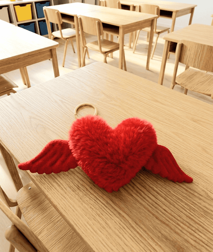 Llavero de corazón rojo con alas visto desde arriba sobre una superficie de madera clara, de fondo puede verse un aula de colegio con mesas y sillas.