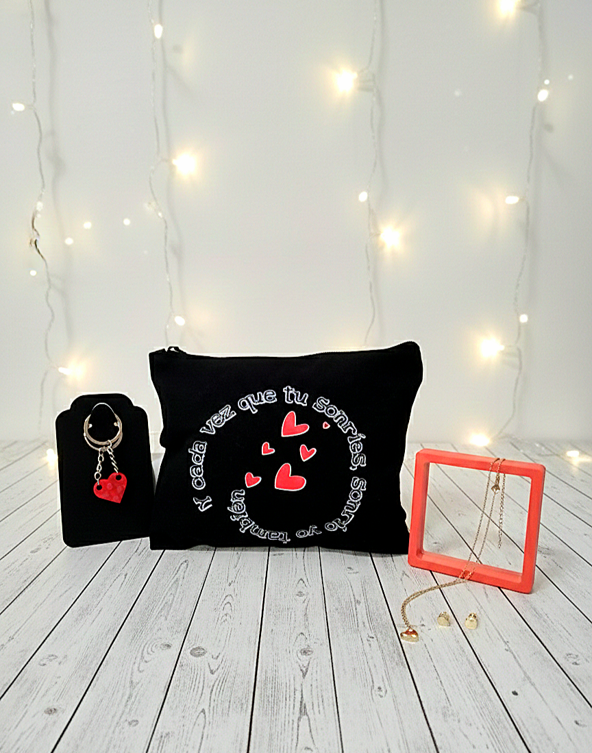 Vista frontal de un kit de regalo que incluye un neceser negro con corazones rojos, un llavero de bloques rojos y joyas doradas frente a un fondo de luces led blancas.