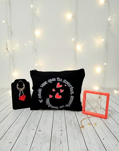 Vista frontal de un kit de regalo que incluye un neceser negro con corazones rojos, un llavero de bloques rojos y joyas doradas frente a un fondo de luces led blancas.