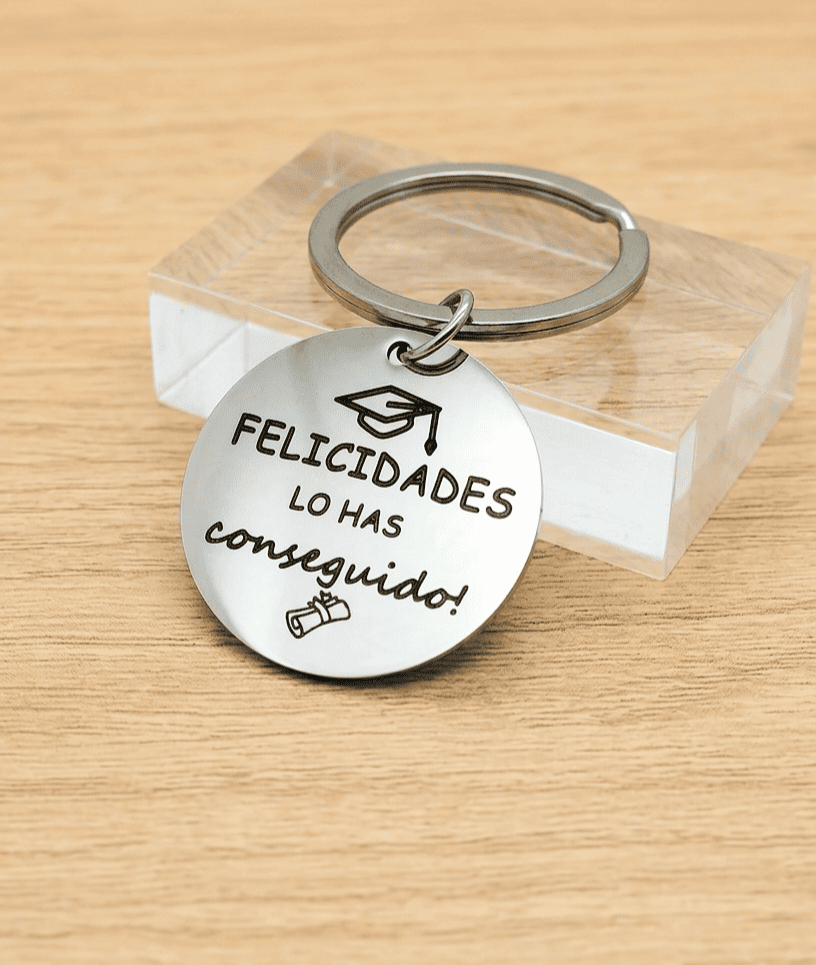 Primer plano de un llavero circular de Acero con la frase "Felicidades lo has conseguido" con un detalle de birrete en la parte superior y un diploma en la parte inferior, el llavero se encuentra apoyado sobre un bloque acrilico transparente de forma rectangutal. 