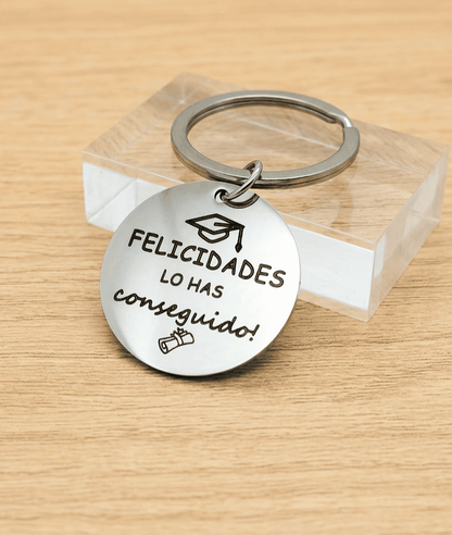  Primer plano de un llavero circular de Acero con la frase "Felicidades lo has conseguido" con un detalle de birrete en la parte superior y un diploma en la parte inferior, el llavero se encuentra apoyado sobre un bloque acrilico transparente de forma rectangutal. 