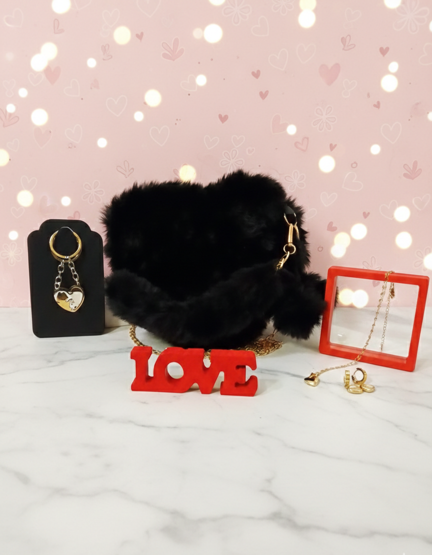Llavero formando un corazon junto a bolso de peluche negro en forma de corazón y set de joyeria con corazones dorados sobre fondo de marmol blaco.