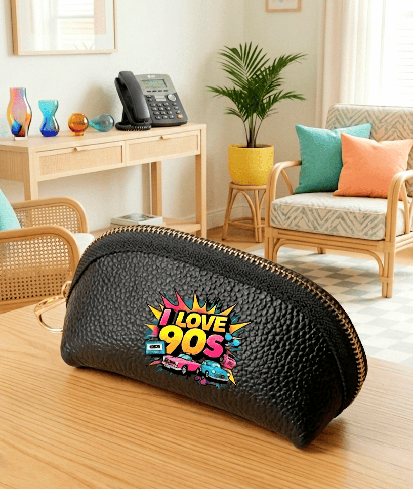 Monedero negro de cuero sintético con cierre de cremallera con un diseño de los 90 compuesto por coches, cintas e casette y la frase "I love 90's" en colores fluor sobre una superficie de madera clara y un fondo de sala de estar con accesorios tipicos de los 90.