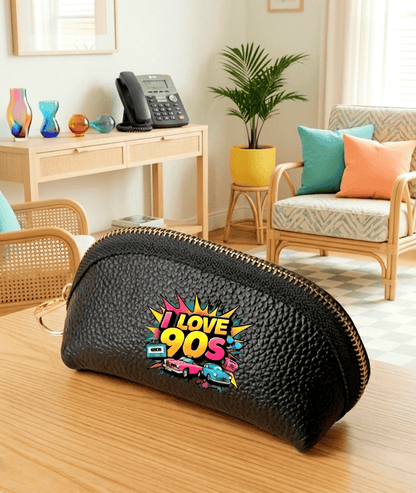 Monedero negro de cuero sintético con cierre de cremallera con un diseño de los 90 compuesto por coches, cintas e casette y la frase "I love 90's" en colores fluor sobre una superficie de madera clara y un fondo de sala de estar con accesorios tipicos de los 90.