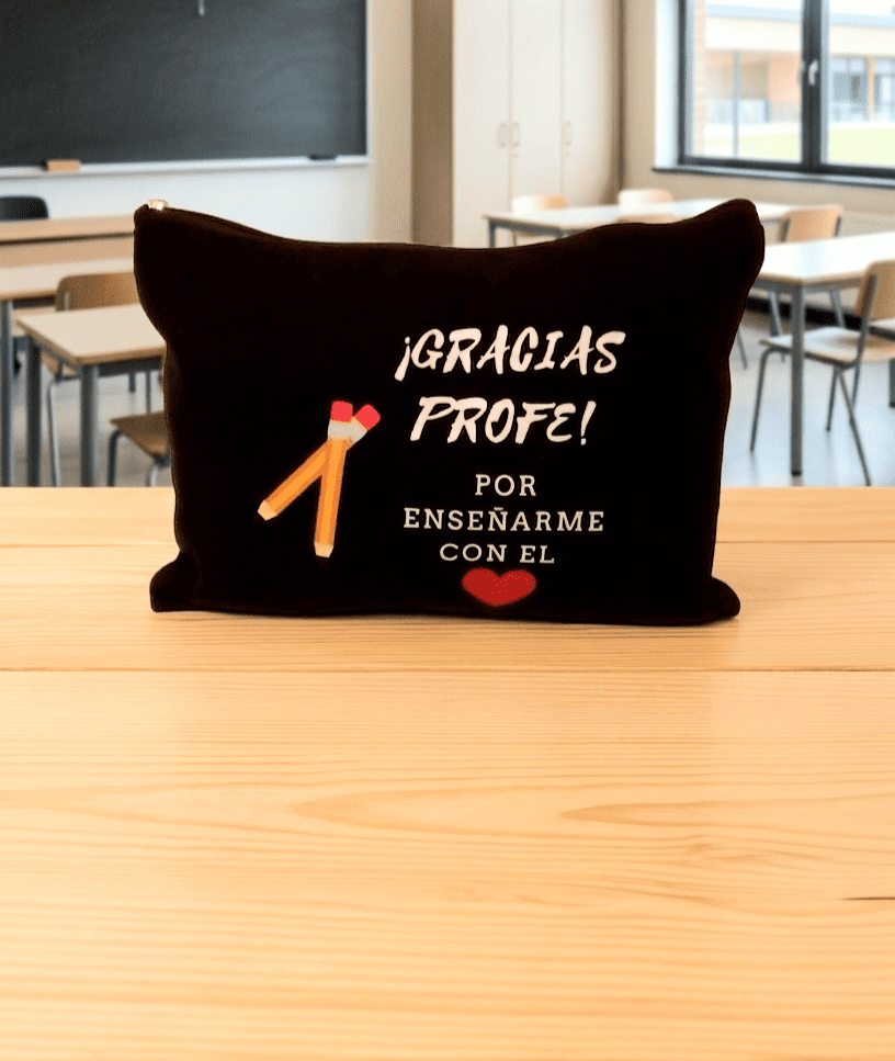Neceser con cierre de cremallera en color negro con la frase de agradecimiento "Gracias Profe por enseñarme con el ❤️" y delalle de dos Lápices cruzados de olor amarillo. Está colocado sobre una superficie de madera clara y de fondo puede verse un aula escolar.