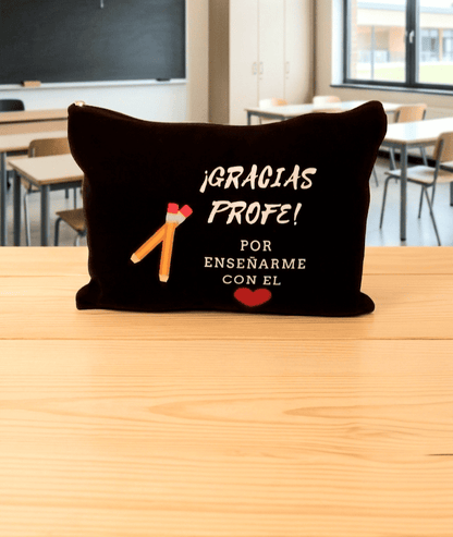 Neceser con cierre de cremallera en color negro con la frase de agradecimiento "Gracias Profe por enseñarme con el ❤️" y delalle de dos Lápices cruzados de olor amarillo. Está colocado sobre una superficie de madera clara y de fondo puede verse un aula escolar.