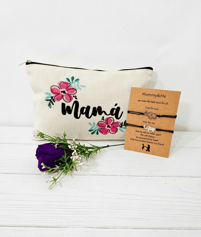 Primer plano de tres articulos de regalo sobre una superficie de madera blanca.A la izquierda un neceser de lona con la palabra "Mama".En el cento una rosa morada artificial. A la derecha una tarjeta kraft con dos pulseras de cuerda negra y dijes de elefantes plateados. La tarjeta lleva el título de" Mommy & me" y contiene un texto en ingles.
