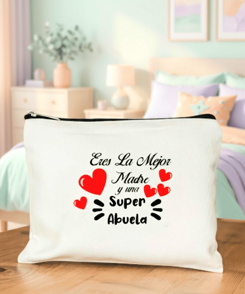 Neceser de lino blanco con la frase "Eres la mejor madre y una superabuela" adornado con corazones rojos sobre superficie de madera y de fondo una cama con colores pastel y muebles y decoración en colores claros.