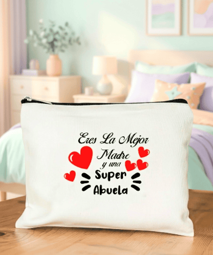 Neceser de lino blanco con la frase "Eres la mejor madre y una superabuela" adornado con corazones rojos sobre superficie de madera y de fondo una cama con colores pastel y muebles y decoración en colores claros.