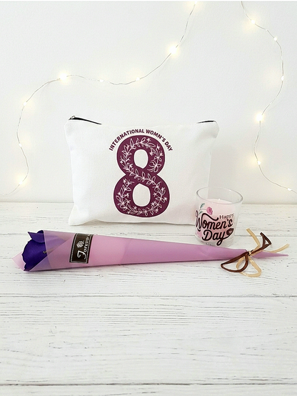 Neceser blanco con numero 8 en color morado junto a una vela rosa con la frase " Happy women's day" y una rosa de color morado con envoltorio en color malva, todo junto sobre una superficie de madera blanca y luces led de fondo.