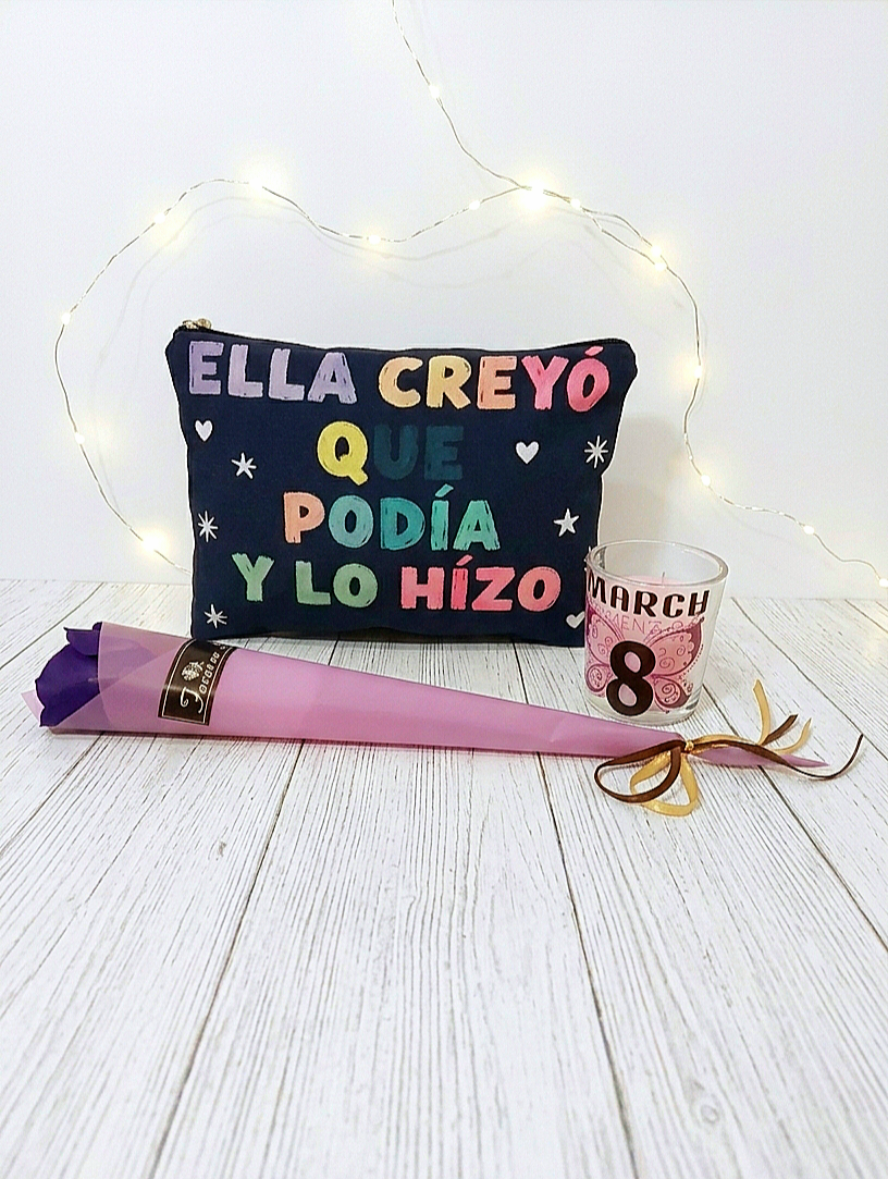Neceser azul oscuro con letras multicolor formando la frase "Ella creyó que podía y lo hizo" junto a una vela con diseño del 8 de Marzo y una rosa morada con envoltorio de color malva sobre superficie de madera blanca y luces led de fondo. 