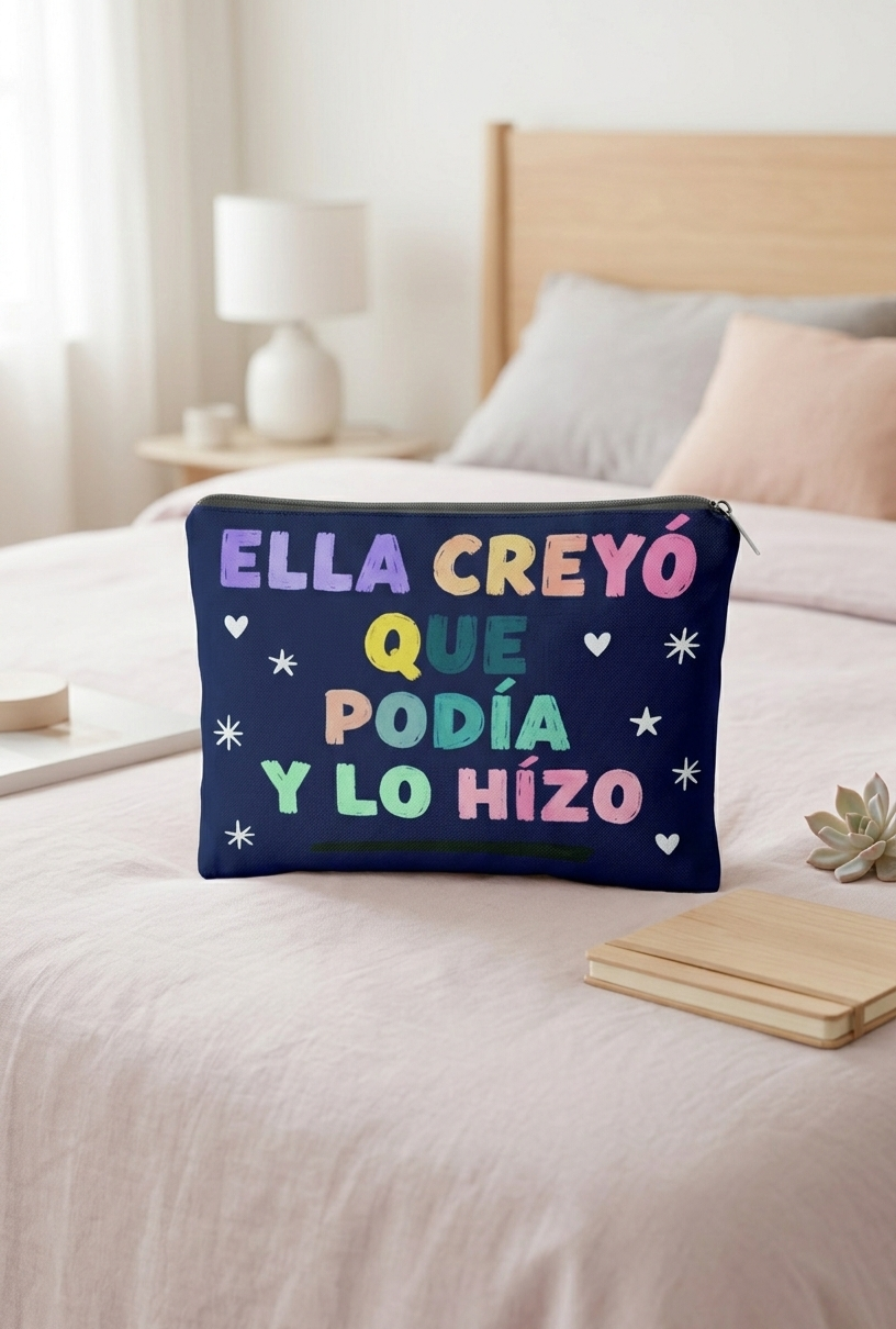 Neceser de color azul oscuro con letras multicolor formando la frase "Ella creyó que podía y lo hizo" sobre una cama de color claro con cojines.