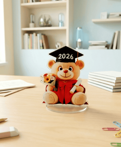 Primer plano de una figura acrilica de osito vestido de Graduación con un birrete negro con el año 2026 en blanco, en una mano sostiene un ramo de flores y en la otra un diploma enrollado, la figura está sobre una base acrilica ovalada colocada sobre un escritorio de madera clara junto a objetos y accesorios escolares con un fondo de habitación juvenil en colores claros.