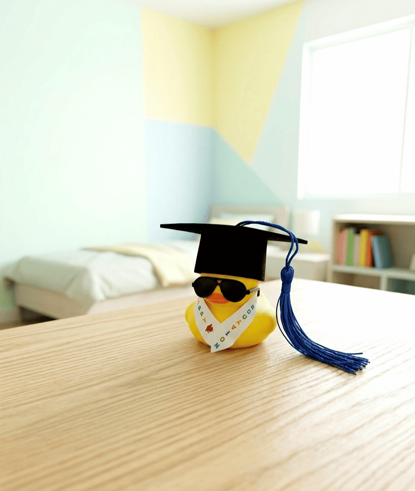 Pato de goma equipado con un birrete negro de borla azul, unas gafas de sol y una estola de graduación con letras de colores sobre una superficie de madera clara y un fondo de habitacion con colores claros.