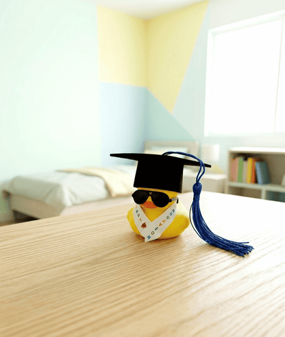 Pato de goma equipado con un birrete negro de borla azul, unas gafas de sol y una estola de graduación con letras de colores sobre una superficie de madera clara y un fondo de habitacion con colores claros.