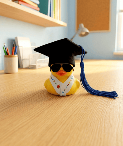 Pato de goma con birrete de borla azul, gafas de sol y estola de graduación sobre una superficie de madera clara y un fondo de habitacion en tonos claros y pastel. 