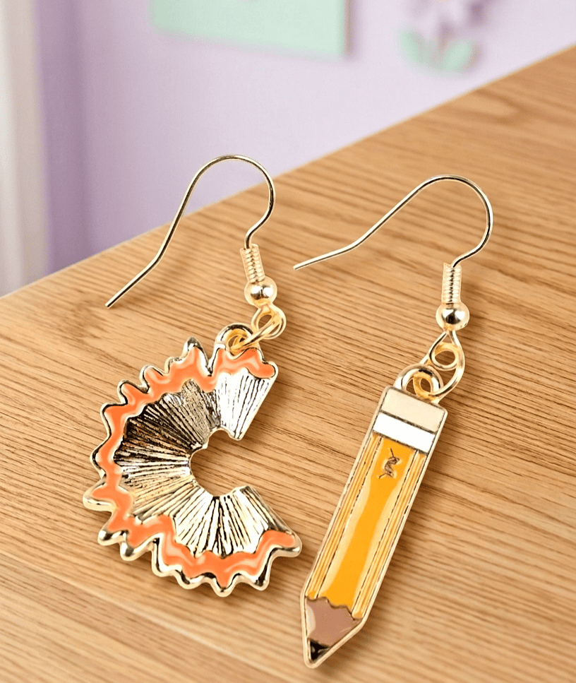 Primer plano de unos pendientes sobre una superficie de madera clara, uno de ellos tiene forma de lapiz de color amarillo esmaltado con detalles dorados y el otro tiene forma de viruta de lapiz en color dorado con detalle de color narsnja esmaltado.