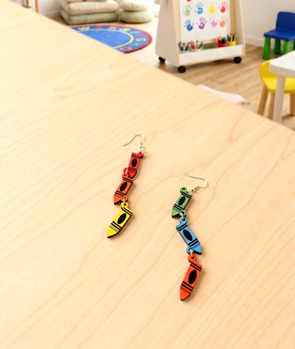 Primer plano de unos pendientes formados por crayones en madera cada uno de un color diferente y unidos entre si con pequeños enganches de metal, se encuentran sobre una superficie de madera clara con un fondo de juguetes y materiales de Escuela Infantil.