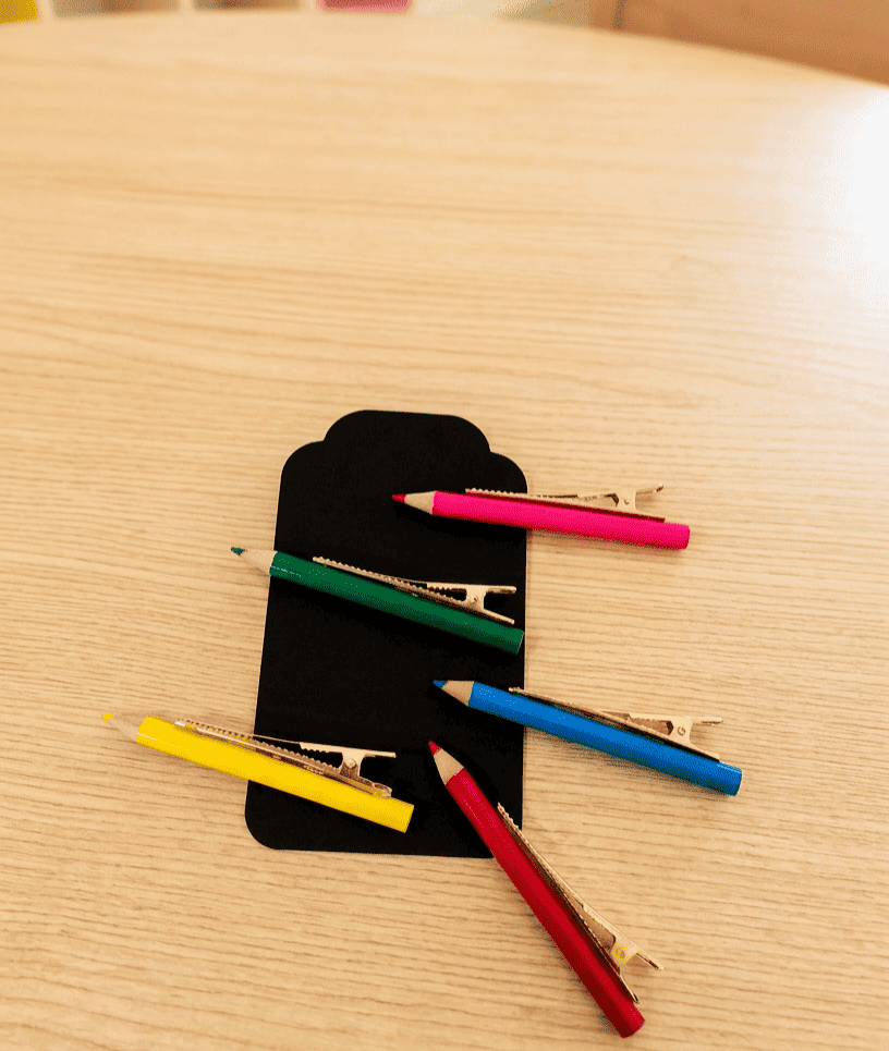 Pinzas con forma de pinturas de colores de madera, compuestas por pinza dorada y pintura de madera cada una de un color diferente, están descolocadas sobre una superficie de madera clara. 