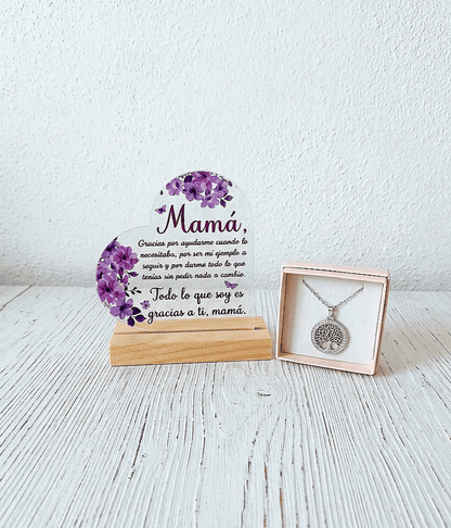 Placa de metacrilato en forma de corazón con un texto emotivo de dia de la madre decorado con flores lilas sobre una base de madera clara junto a una caja con un colgante con Arbol de la vida plateado en Acero inoxidable, todo sobre una superficie de madera blanca. 