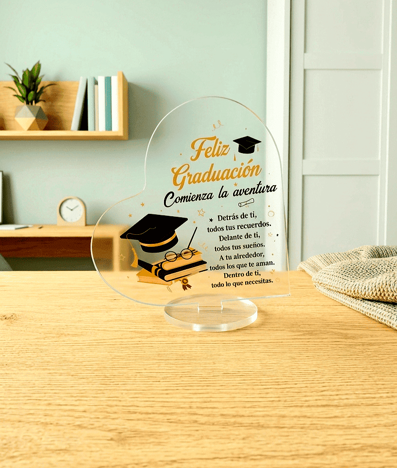 Placa acrilica transparente en forma de corazón con un texto emotivo en español y motivos de graduación sobre una base redonda giratoria. De fondo se puede ver una habitación juvenil de colores claros.