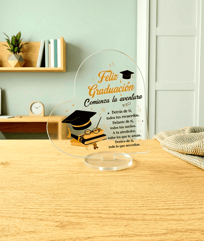 Placa acrilica transparente en forma de corazón con un texto emotivo en español y motivos de graduación sobre una base redonda giratoria. De fondo se puede ver una habitación juvenil de colores claros.