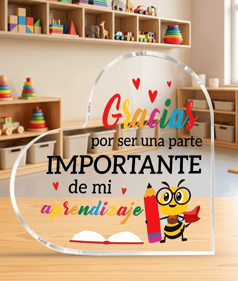 Placa acrilica en forma de corazón con un mensaje de agradecimiento " Gracias por ser una parte Importante de mi aprendizaje" con detalle de abeja con lapiz y un libro abierto, sobre una superficie de madera clara con un fondo de aula de Escuela infantil. 