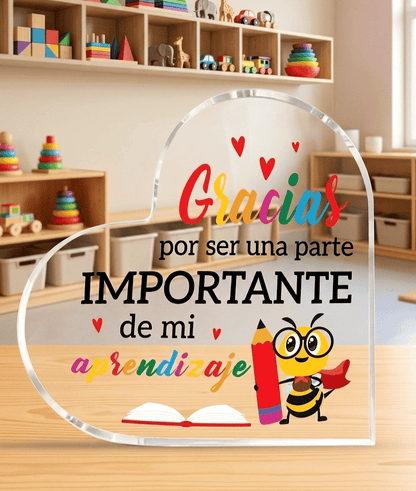 Placa acrilica en forma de corazón con un mensaje de agradecimiento " Gracias por ser una parte Importante de mi aprendizaje" con detalle de abeja con lapiz y un libro abierto, sobre una superficie de madera clara con un fondo de aula de Escuela infantil. 