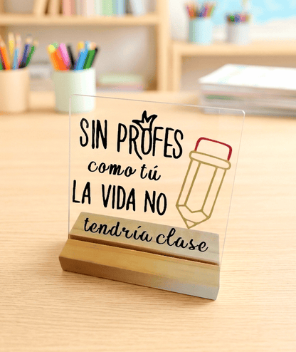 Primer plano de una Placa Acrilica con la frase "SIN PROFES COMO TU LA VIDA NO TENDRÍA CLASE" acompañada del dibujo de un lapicero en una base de madera clara sobre un escritorio de madera con articulos y detalles escolares de fondo.