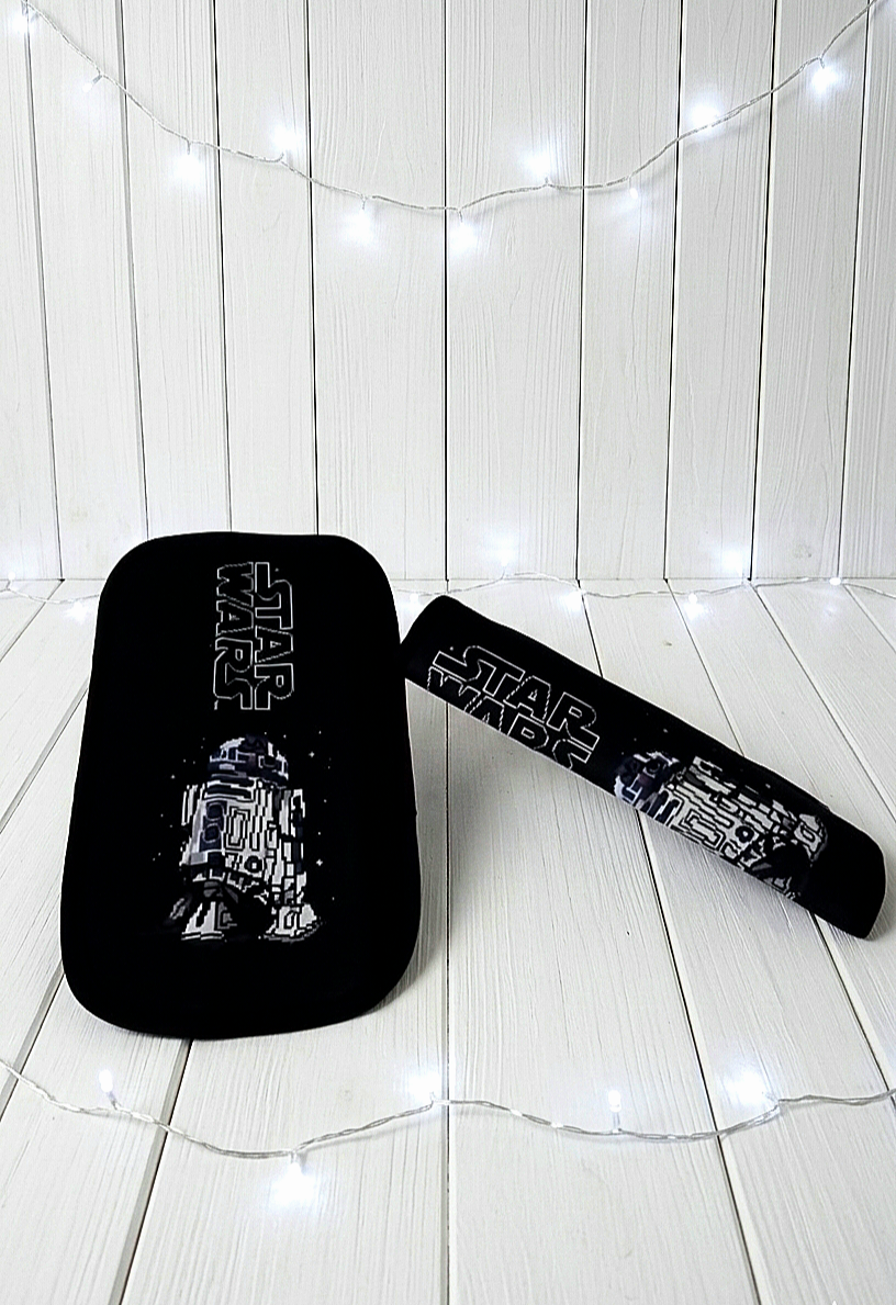 Protector de cinturón de seguridad extendido con diseño de R2 D2 , junto a otro plegado sobre superficie de madera blanca rodeado de luces led.
