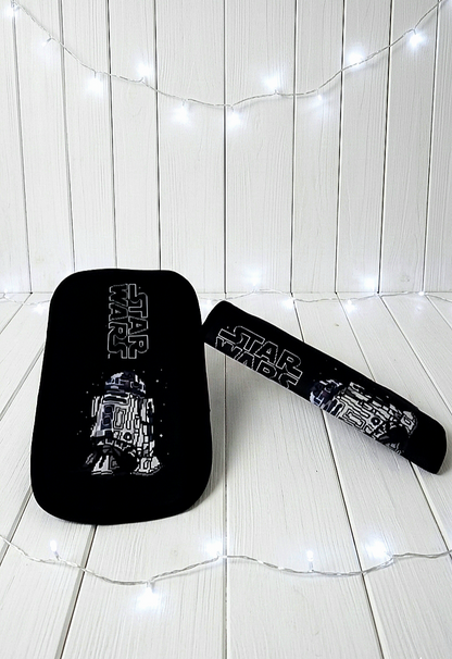 Protector de cinturón de seguridad extendido con diseño de R2 D2 , junto a otro plegado sobre superficie de madera blanca rodeado de luces led.