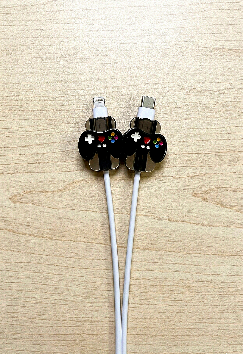 Dos protectores de cable de carga en forma de mini controles de videojuegos negros con detalles de coloes sbre fondo de madera clara