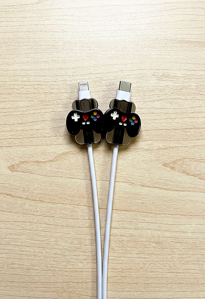 Dos protectores de cable de carga en forma de mini controles de videojuegos negros con detalles de coloes sbre fondo de madera clara