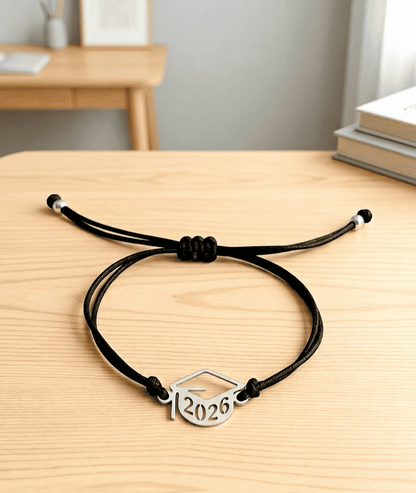 Pulsera de cordón negro ajustable con un dije metálico de birrete con el año 2026 sobre una superficie de madera clara con un fondo de habitación con muebles de colores claros.