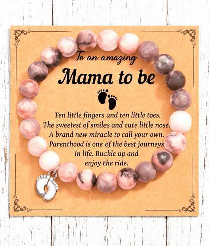 Primer plano de una pulsera de cuentas en color pastel con dije de pies de bebé en color plateado sobre una tarjeta de papel kraft con un texto emotivo en inglés para fururas mamas. 