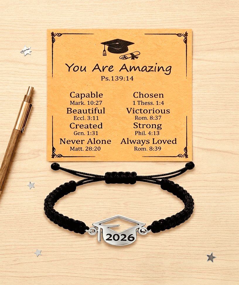 Pulsera negra ajustable con detalle metalico en forma de birrete con el año 2026 junto a tarjeta kraft con un texto inspirador en inglés, sobre una superficie de escritorio de madera clara, en la parte izquierda de la imagen hay un boligrafo y sobre la superficie pequeñas estrellitas plateadas.