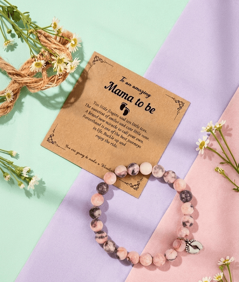 Pulsera de cuentas en colores pastel con dije de pies de bebé en color plateado junto a una tarjeta de papel Kraft con texto en ingles de día de la madre sobre un fondo en colores pastel.