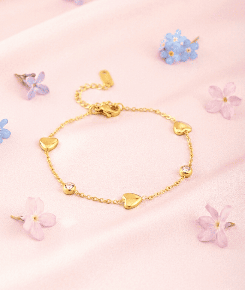 Pulsera dorada en Acero Inoxidable compuesta por corazones e incrustaciones de cristales brillantes, vista desde arriba sobre un fondo de satén rosa pastel con pequeñas flores silvestres alrededor. 
