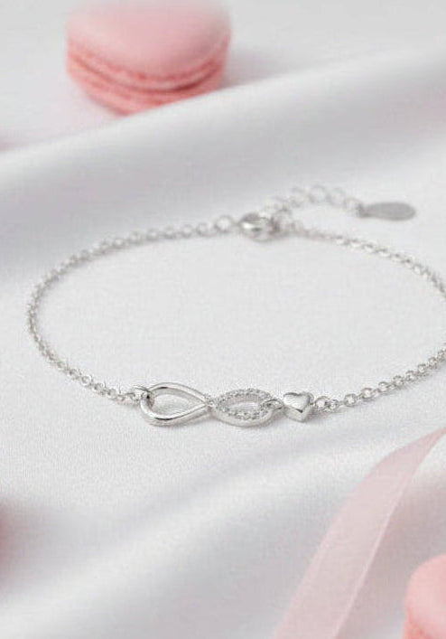 Pulsera de plata con simbolo de infinito y pequeño corazón sobre fondo de seda blanca.