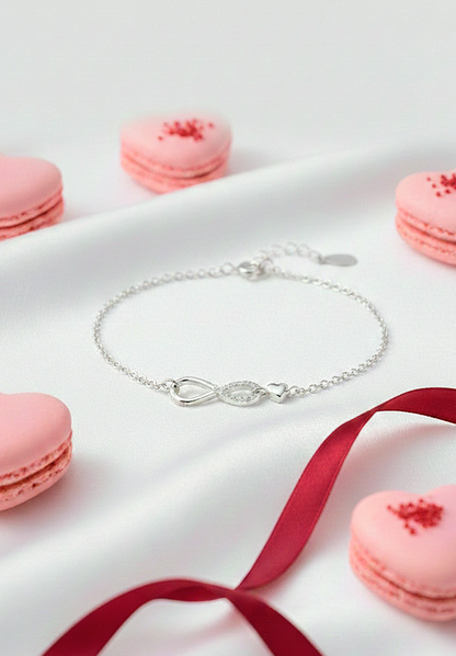 Pulsera de plata con simbolo de Infinito y pequeño corazón sobre fondo de seda blanca.
