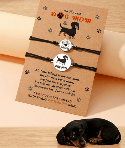 Set de pulseras de cordón negro encerado con dijes de motivos de perro y huella de patita, presentadas en tarjeta de papel kraft con mensaje en inglés. En la parte inferior de la imagen se puede ver a un perrito negro tumbado.
