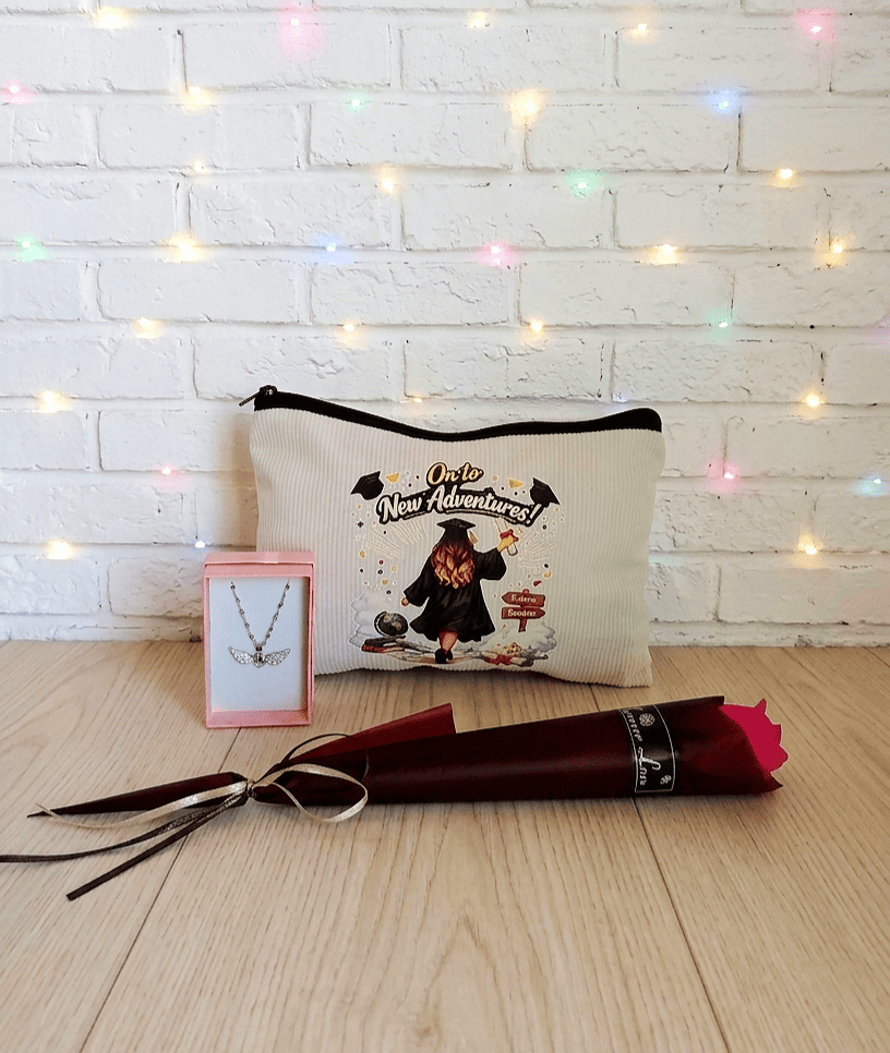 Set de regalos de Graduación sobre tarima de madera clara, puede verse una caja de joyeria y en su interior un colgante de Acero Inoxidable de corazón con alas brillantes, en el centro puede verse un neceser con un diseño e ilustración de Graduación y en la parte superior la frase: "On to new adventures", en la parte inferior de la imagen puede verse una Rosa Eterna con envoltorio de papel satinado de color Borgoña.