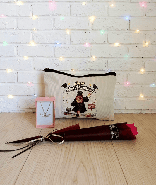 Set de regalos de Graduación sobre tarima de madera clara, puede verse una caja de joyeria y en su interior un colgante de Acero Inoxidable de corazón con alas brillantes, en el centro puede verse un neceser con un diseño e ilustración de Graduación y en la parte superior la frase: "On to new adventures", en la parte inferior de la imagen puede verse una Rosa Eterna con envoltorio de papel satinado de color Borgoña.