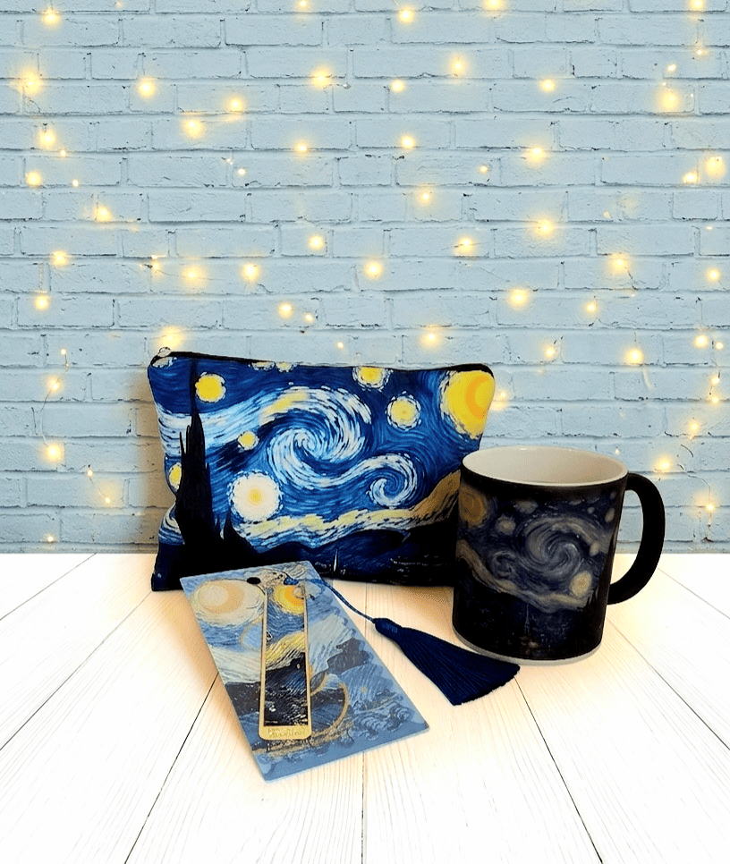 Conjunto de regalos con estética de cuadro "La noche estrellada" de Van Gogh,  compuesto por un neceser con diseño de "La noche estrellada" una taza de ceramica termosensible con diseño del mismo cuadro y un marcapaginas con relieve dorado y diseño de "La noche estrellada" con adorno azul cobalto presentado en una tarjeta con los mismos colores y diseño que el resto de los artículos, todo sobre una superficie de madera blanca con pared azulada y luces Led. 