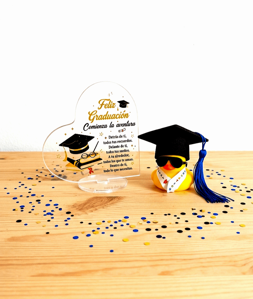 Conjunto de articulos de regalo de graduación compuesto por una placa acrilica con forma de corazón y un texto emotivo de graduación junto a un pato de goma con birrete negro, gafas de sol y una estola con letras de colores, todo sobre un fondo de madera clara con confeti de colores metalizados.