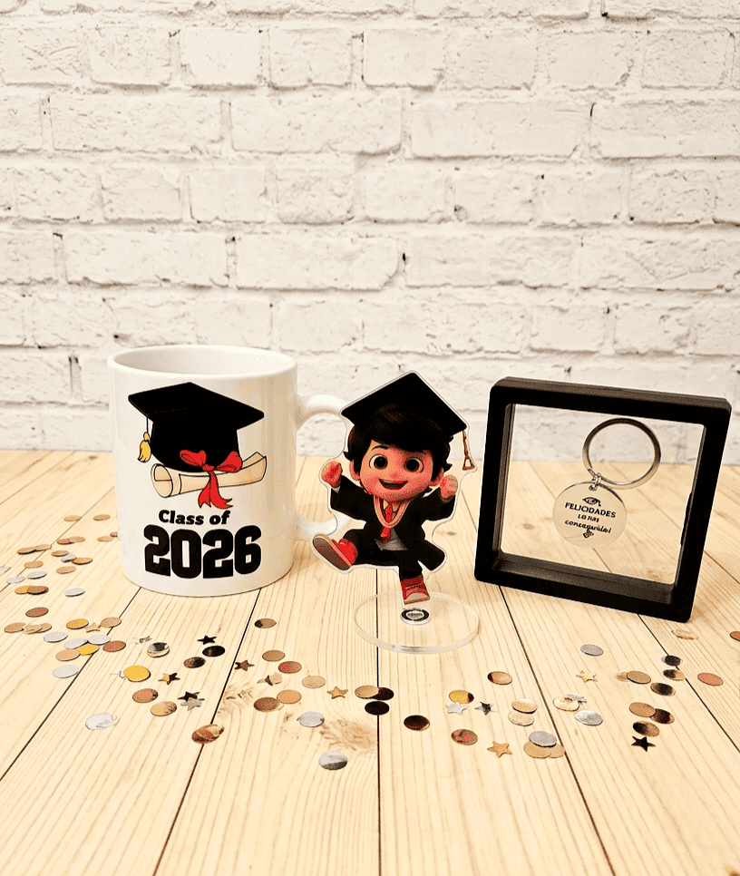 Conjunto de regalos de graduación compuesto por una taza con diseño de birrete con diploma de graduación con la frase "Class of 2026" a su lado una figura acrilica con base giratoria vestido con una toga y un birrete con expresión de alegría, por último un llavero de acero con la frase grabada "Felicidades lo has conseguido" presentado en caja de exhibición con marco negro, todo sobre una superficie de madera clara con confeti metalizado alrededor.