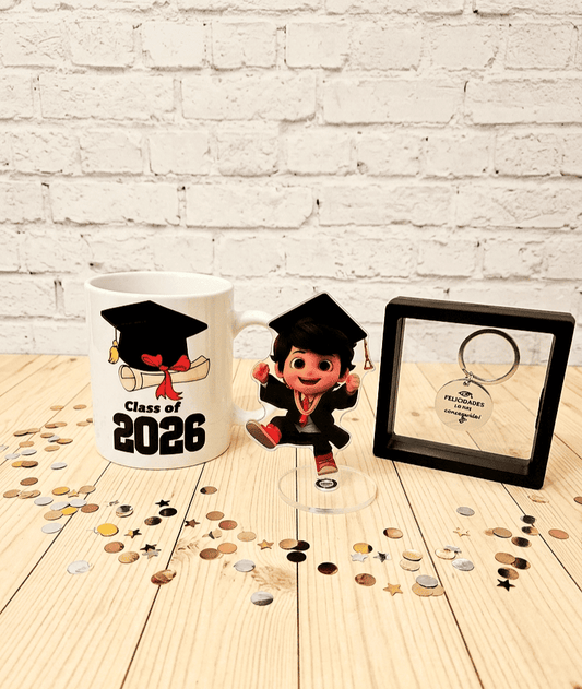 Conjunto de regalos de graduación compuesto por una taza con diseño de birrete con diploma de graduación con la frase "Class of 2026" a su lado una figura acrilica con base giratoria vestido con una toga y un birrete con expresión de alegría, por último un llavero de acero con la frase grabada "Felicidades lo has conseguido" presentado en caja de exhibición con marco negro, todo sobre una superficie de madera clara con confeti metalizado alrededor.