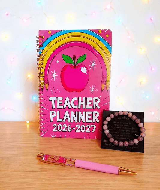 Conjunto de regalos para profesora sobre una superficie de madera clara y pared blanca con luces led, formado por una agenda planificadora de color fucsia con diseño Retro-Pop, a su lado se encuentra una pulsera de cuentas en tonos rosados presentada en tarjeta negra con dedicatoria de letras en dorado, por ultimo en el centro se puede ver un boligrafo de color rosa en la parte inferior y acrilico con flores fucsias en la parte superior.