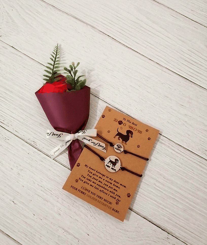 Conjunto de regalos compuesto por un set de pulseras presentadas en una tarjeta de papel kraft con un mensaje en inglés, junto a una Rosa Eterna de jabón con envoltorio de papel satinado de color burdeos, todo sobe una superficie de madera blanca. 