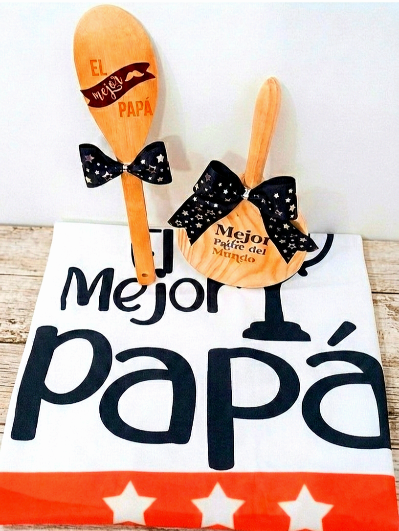 Conjunto de articulos de cocina compuesto por una cuchara de madera grabada, una tabla de cortar redonda, ambas con lazos negros de estrellas plaeadas, apoyadas sobre el delantal de cocina plegado que muestra la frase "EL MEJOR PAPÁ " y una franja naranja con estrellas blancas